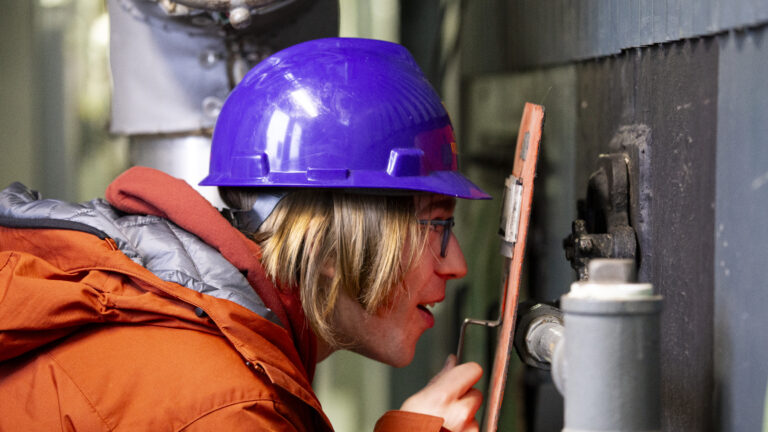 Student wearing a purple hard hat, using equipment to examine a lit flame