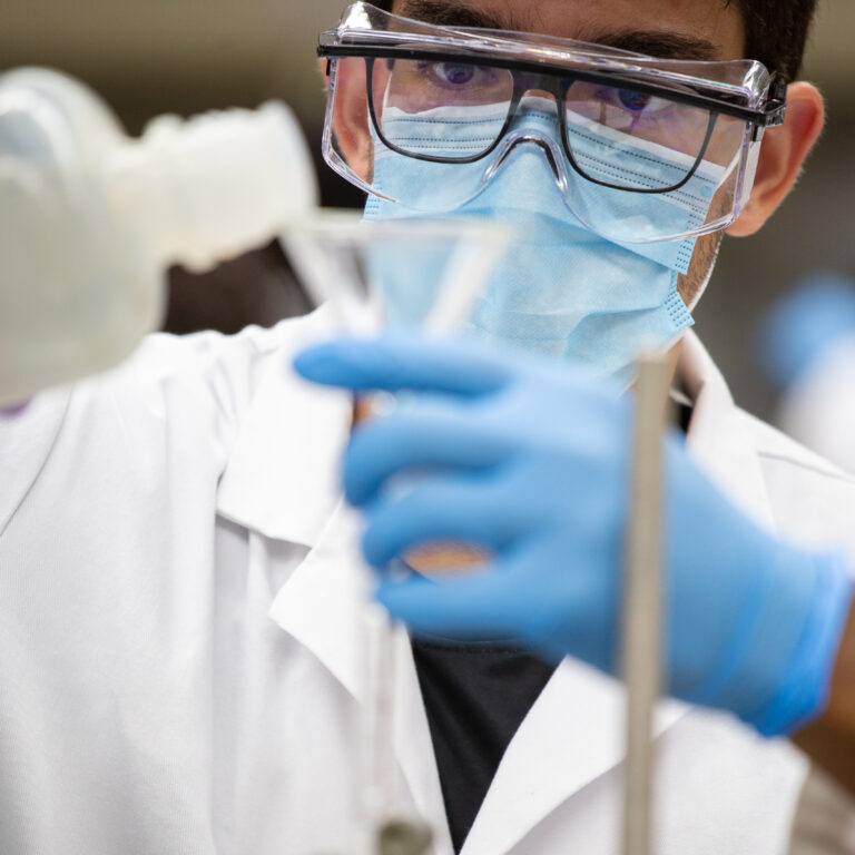 A student with lab coat and protective eyewear pouring liquid into a funnel in a science lab