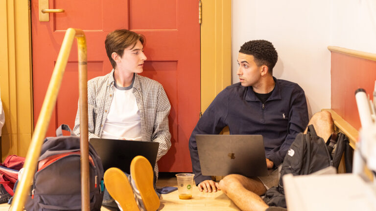 Two students sit leaning against the wall having a conversation in Goodrich Hall
