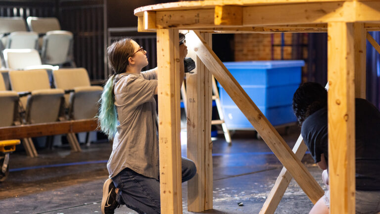 A student with a power drill working on wooden stage equipment for a performance