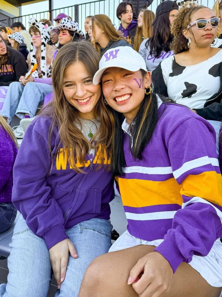 Two students wearing purple and gold Williams shirts smiling in the stands of a football game.
