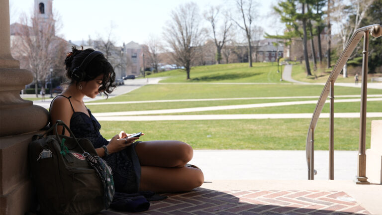 A student wearing headphones and typing on her phone sits in a sunny patch on the Hopkins Hall stairs
