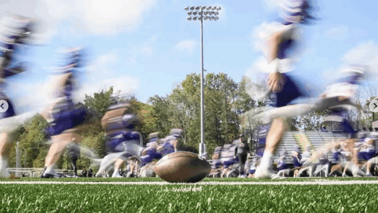 A still football laying on a football field while a group of blurry football players in uniform run by