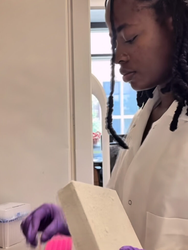 A student wearing a lab coat and gloves working with materials in a lab