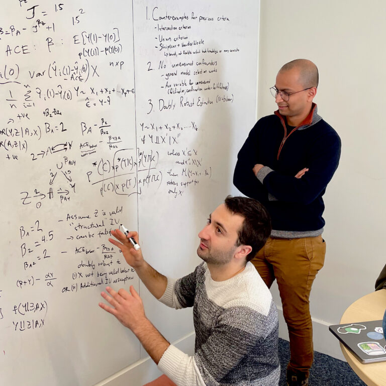 A student discusses equations on a whiteboard with two other students and a professor