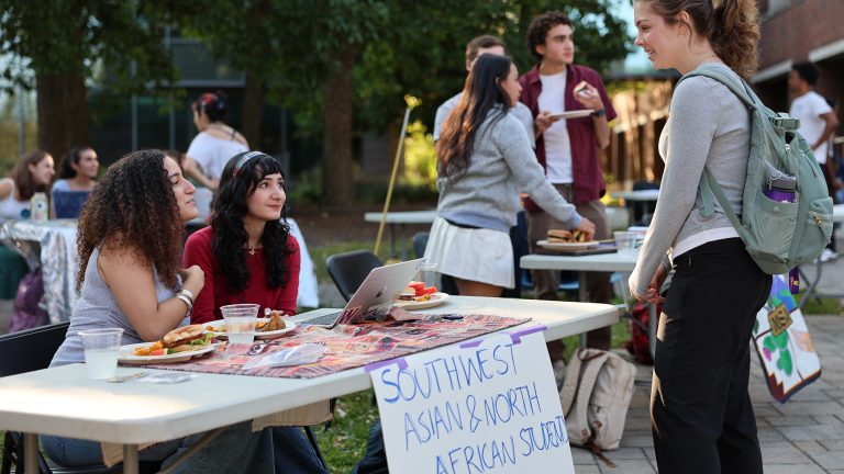 Two students sitting at a table for the Southwest Asian & North African Student Group at a tabling event talking to a student who approached the table