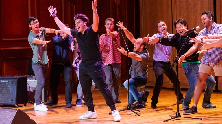 A student group performing on stage and pointing to a featured performer front and center of the group