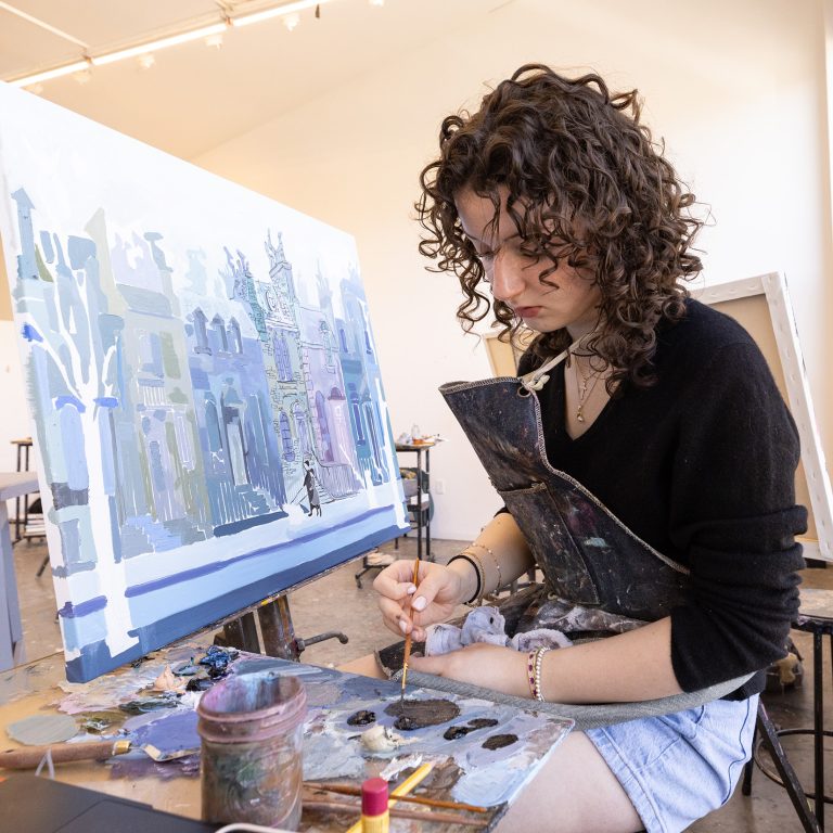 A student adding more paint to their brush as they paint on a canvas
