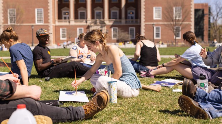 A group of students sitting on the grass in the quad while painting on notepads and notebooks