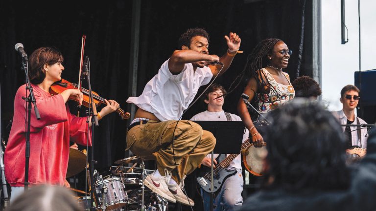 A singer jumping while singing into a microphone with a band playing behind them