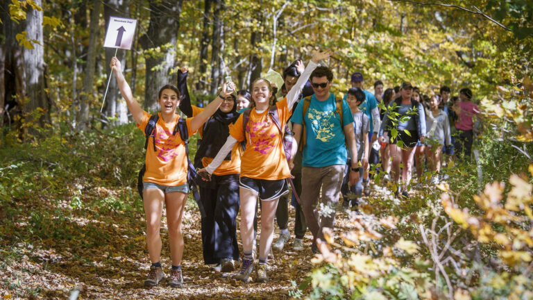 Students hold up their hands and cheer as they hike through the woods.